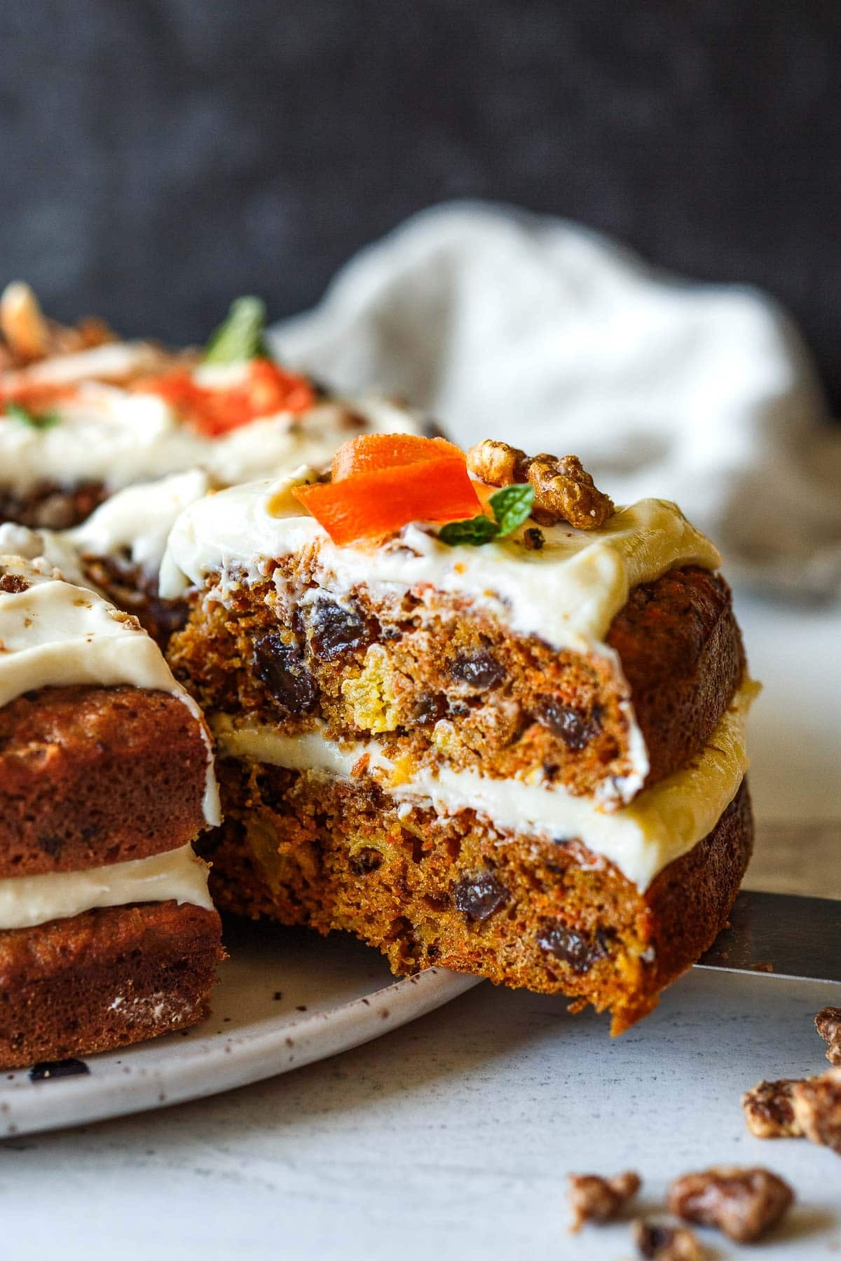 carrot cake slice with cream cheese frosting, with walnuts and raisins, with a garnish of carrot, mint, and walnut. 