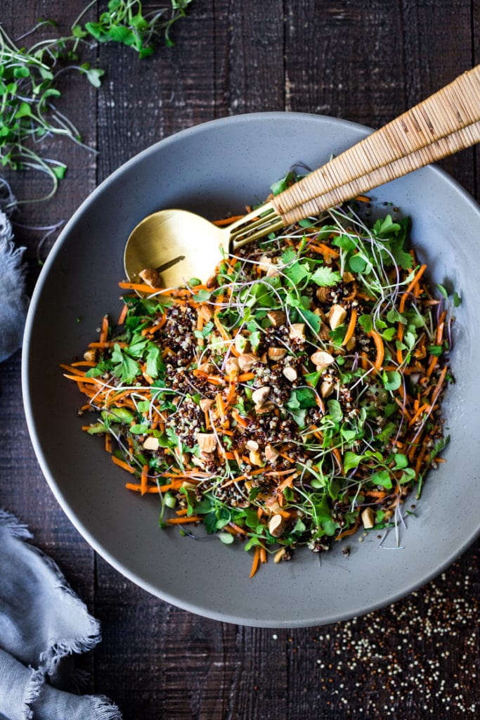 salad bowl with carrot quinoa salad, one of our healthy carrot recipes, with fresh herbs, microgreens, nuts, shredded carrots, and tri-color quinoa with serving utensils.