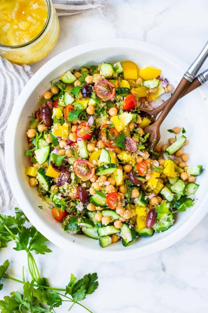 bowl of quinoa chickpea salad with colorful veggies, cucumber, cherry tomatoes, chickpeas, fresh herbs, olives, with wood serving utensils. 