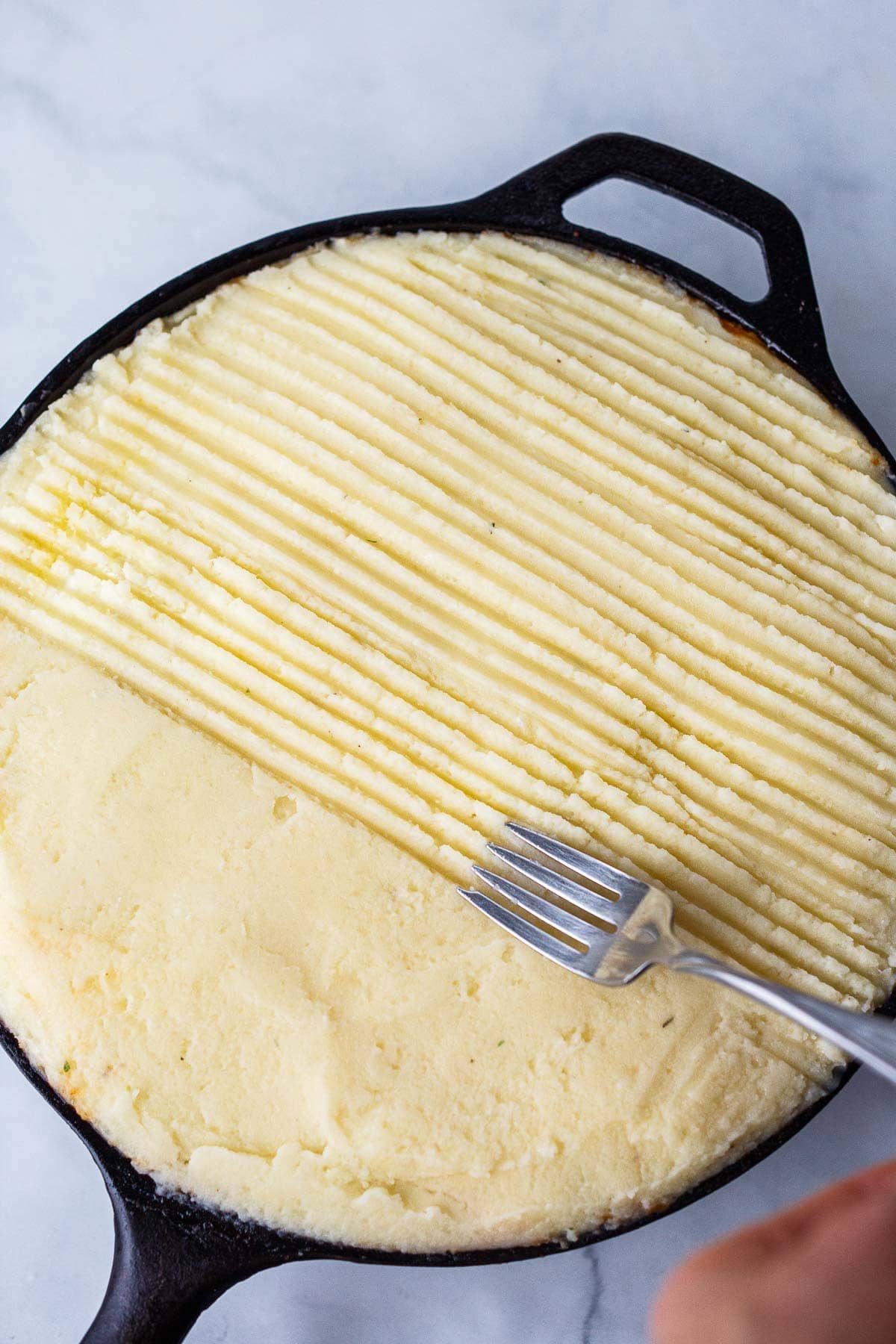 fork making grooves across mashed potato topping on cottage pie in cast iron skillet.
