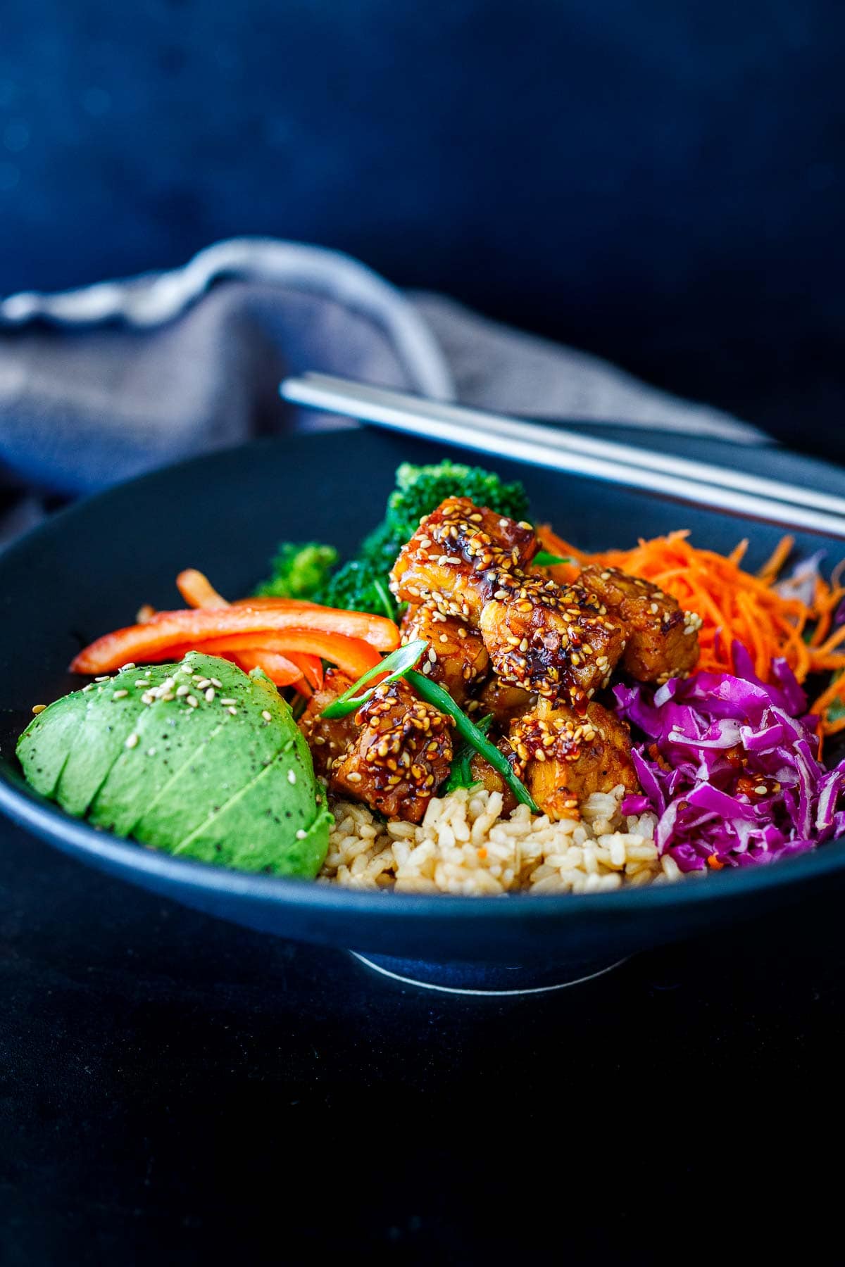 crispy sesame tempeh bowl with rice, avocado, purple cabbage, carrots, peppers, and broccoli with chopsticks on the bowl. 