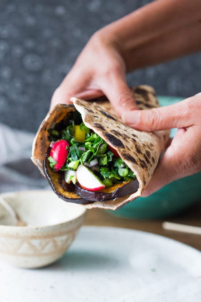 hands holding a Middle Eastern eggplant wrap with greens, radish, and veggies. 