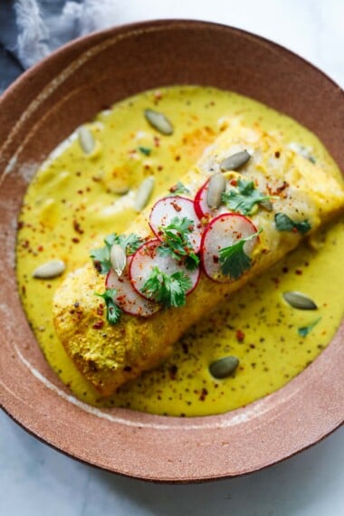 shrimp enchiladas, one of our most popular Mexican recipes, in a yellow sauce garnished with pumpkin seeds, radishes, and cilantro.