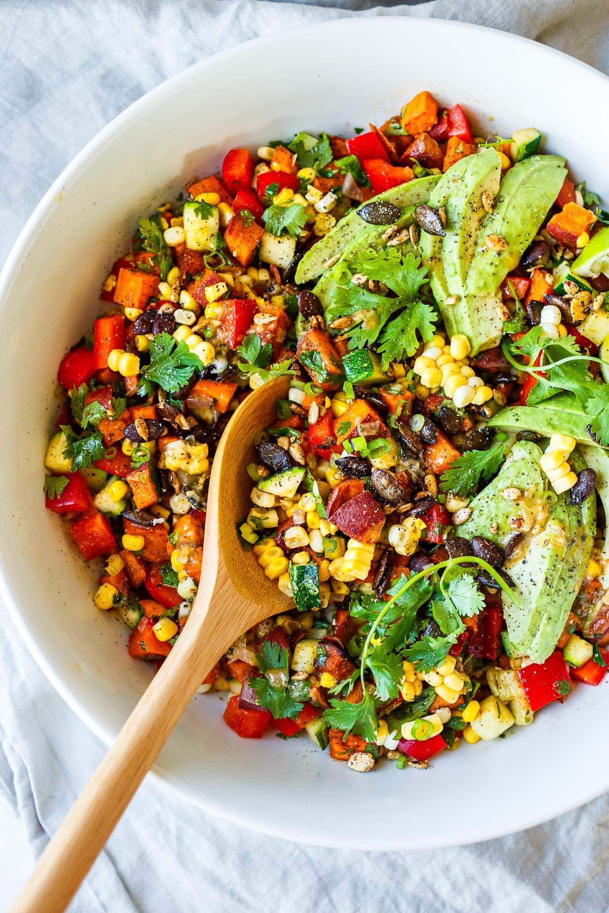 big bowl with sweet potato salad with corn, beans, sweet potato, peppers, green onion, herbs, and sliced avocado with big wood spoon.