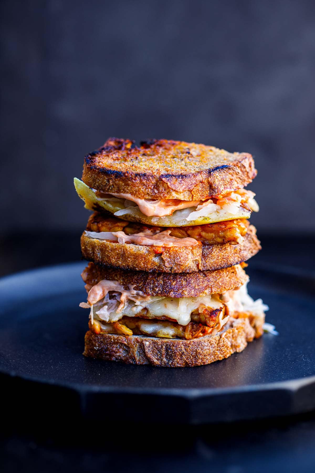 two tempeh reuben sandwiches stacked on top of each other with melty cheese, sauerkraut, and Reuben sauce.