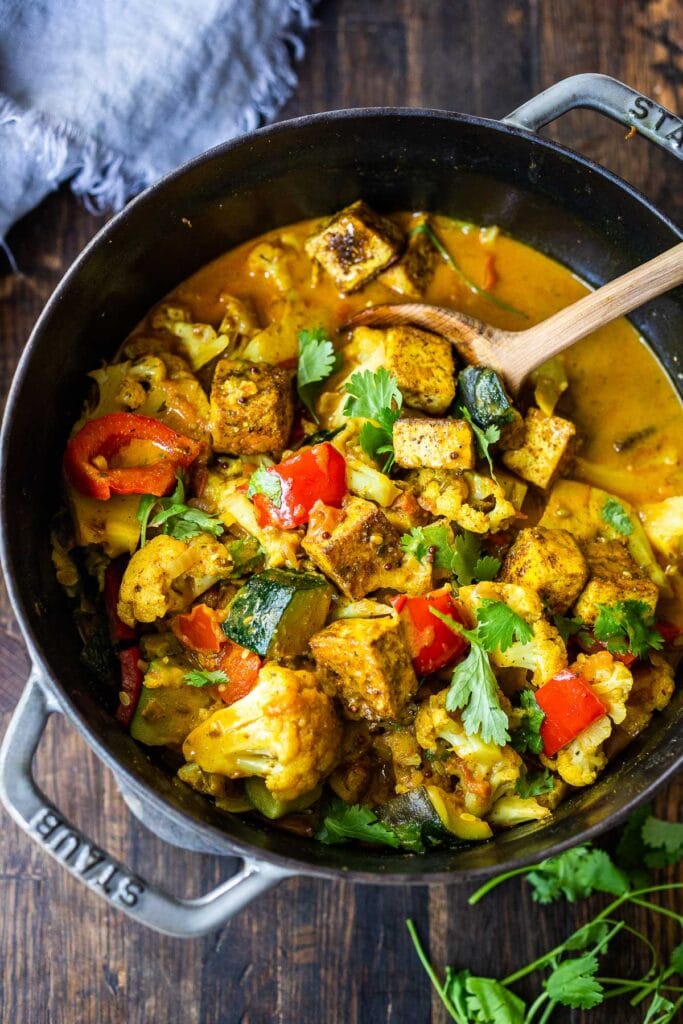 vegan tikka masala in skillet with tofu, cauliflower, peppers, and zucchini with wood spoon, garnished with cilantro.