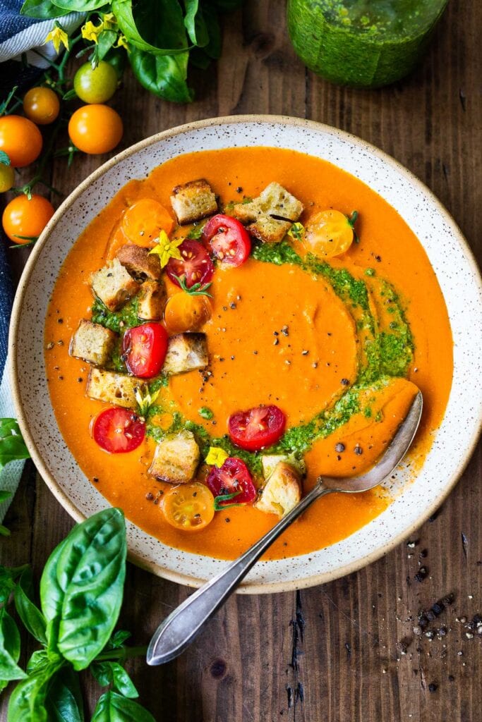 bowl of tomato soup with a swirl of pesto and topped with croutons and fresh cherry tomatoes with a spoon in the bowl. 