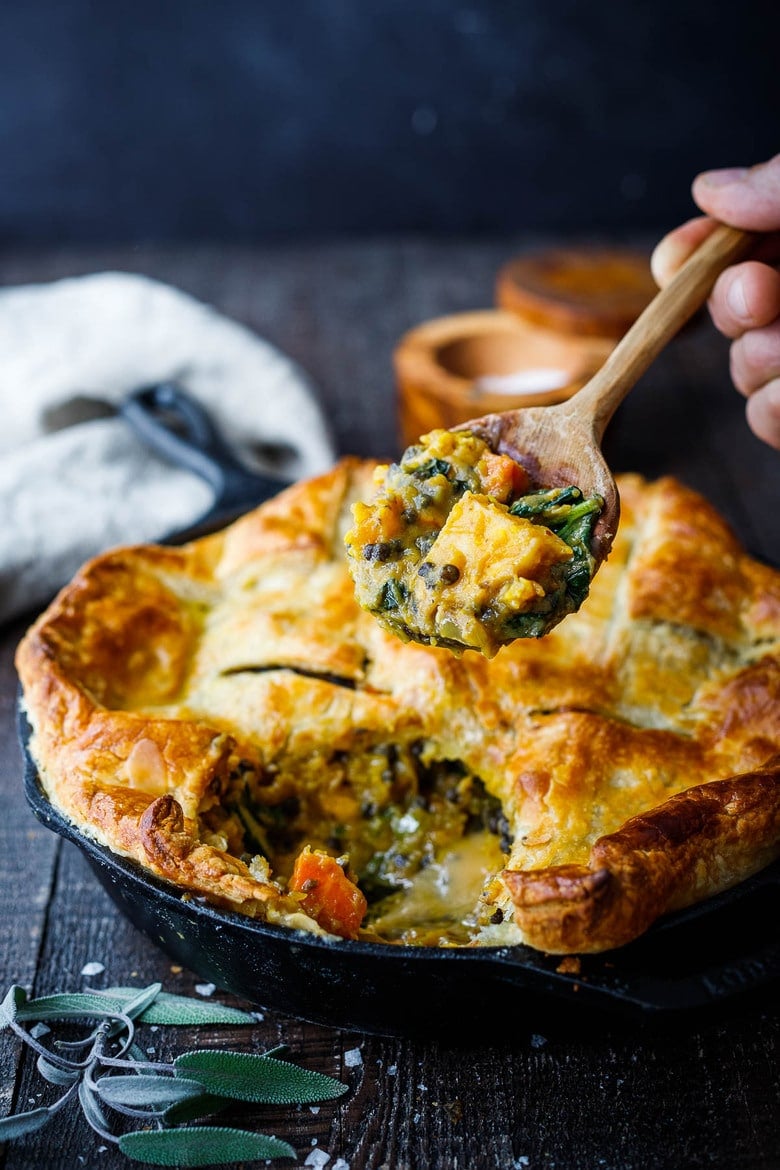 cast iron skillet with lentil pot pie with puff pastry crust and wood spoon holding up spoonful of veggies.